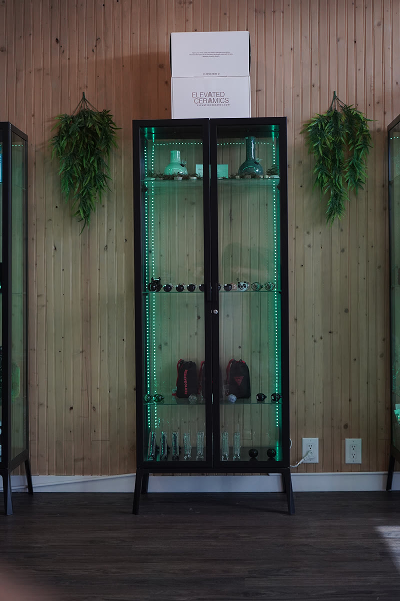 A black-framed glass cabinet with green LED strips on a cedar-panel wall, glass and accessories inside.